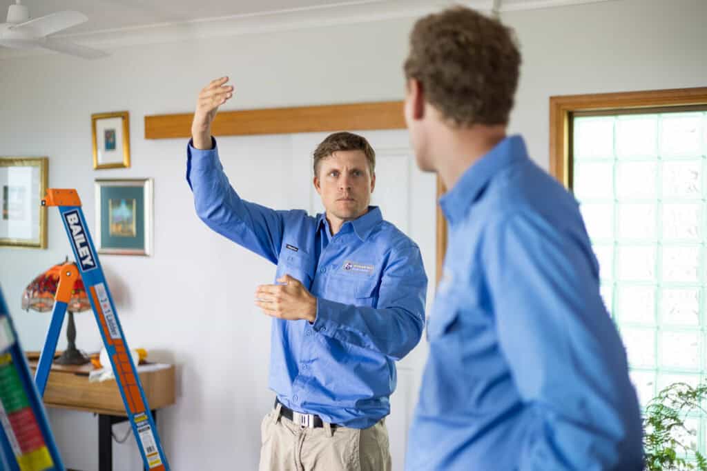 A Caucasian man in a blue work shirt and khaki pants is explaining something to another man, gesturing with his hands. A "Bailey" brand orange ladder is visible on the left side of the frame, indicating work being done indoors.