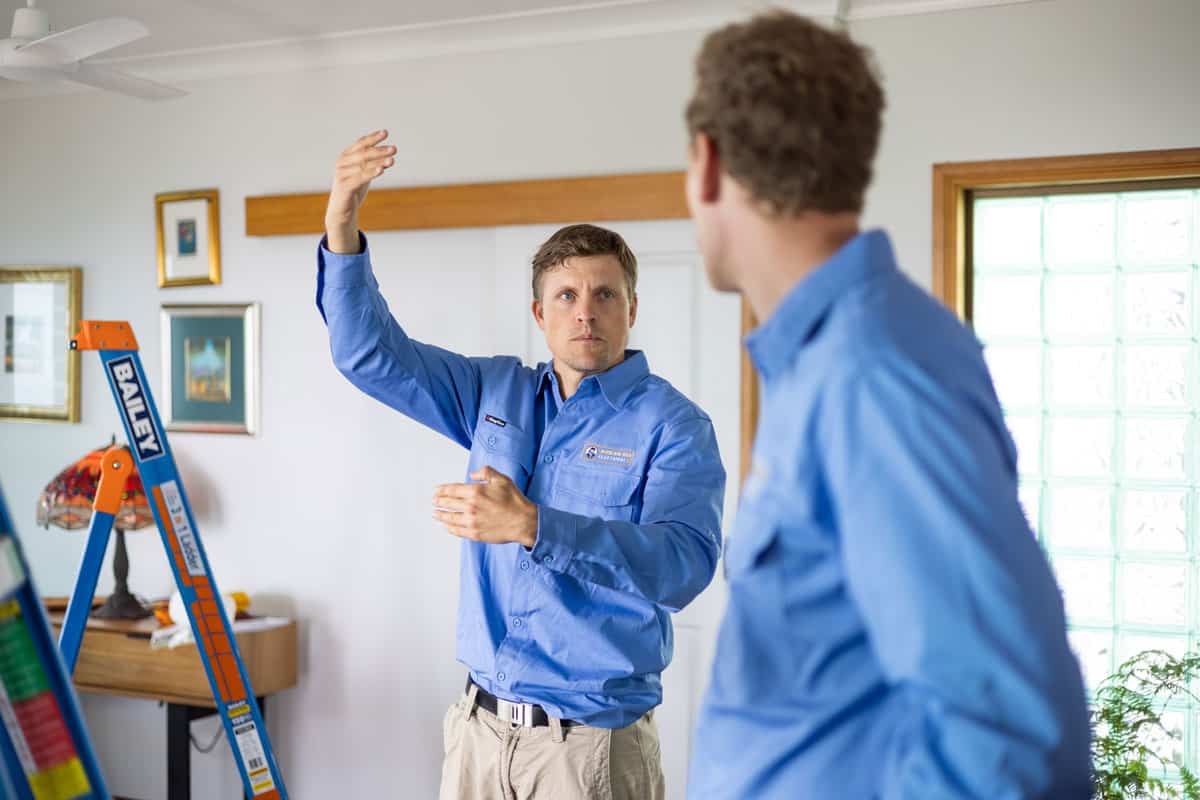 A Caucasian man in a blue work shirt and khaki pants is explaining something to another man, gesturing with his hands. A "Bailey" brand orange ladder is visible on the left side of the frame, indicating work being done indoors.